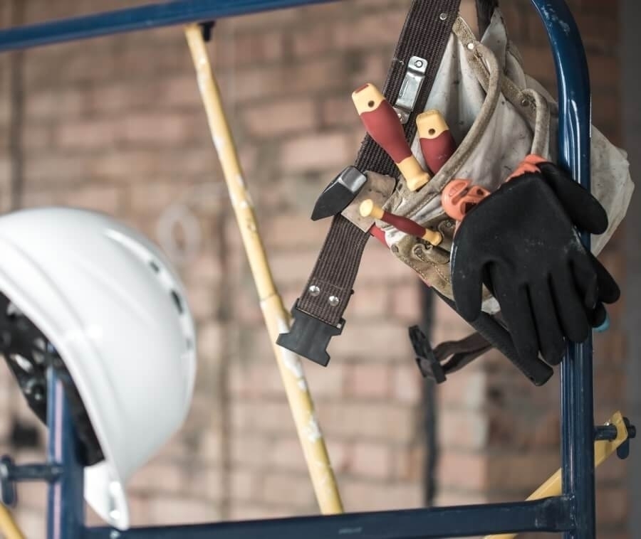 Hard hat, tool belt, gloves, and screwdrivers hanging on scaffolding at construction site.