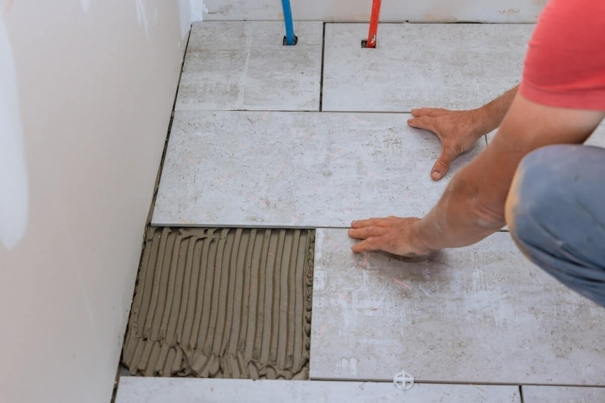 Hands laying large format gray bathroom floor tiles with thinset mortar.